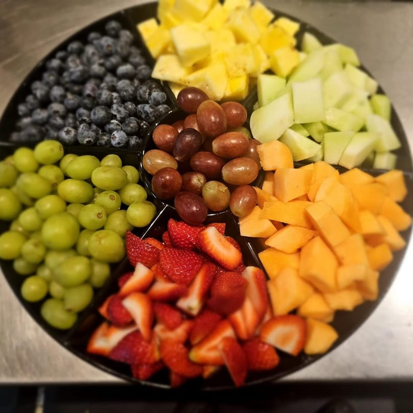 A black platter filled with sections of fresh blueberries, pineapple, honeydew, cantaloupe, strawberries, green grapes, and red grapes.