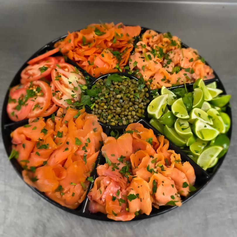 A platter of smoked salmon, sliced tomatoes, capers, and lime wedges, garnished with parsley.