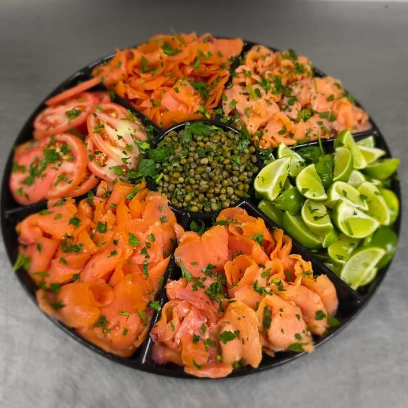 A catering platter with smoked salmon, sliced tomatoes, capers, and lime wedges, garnished with parsley.